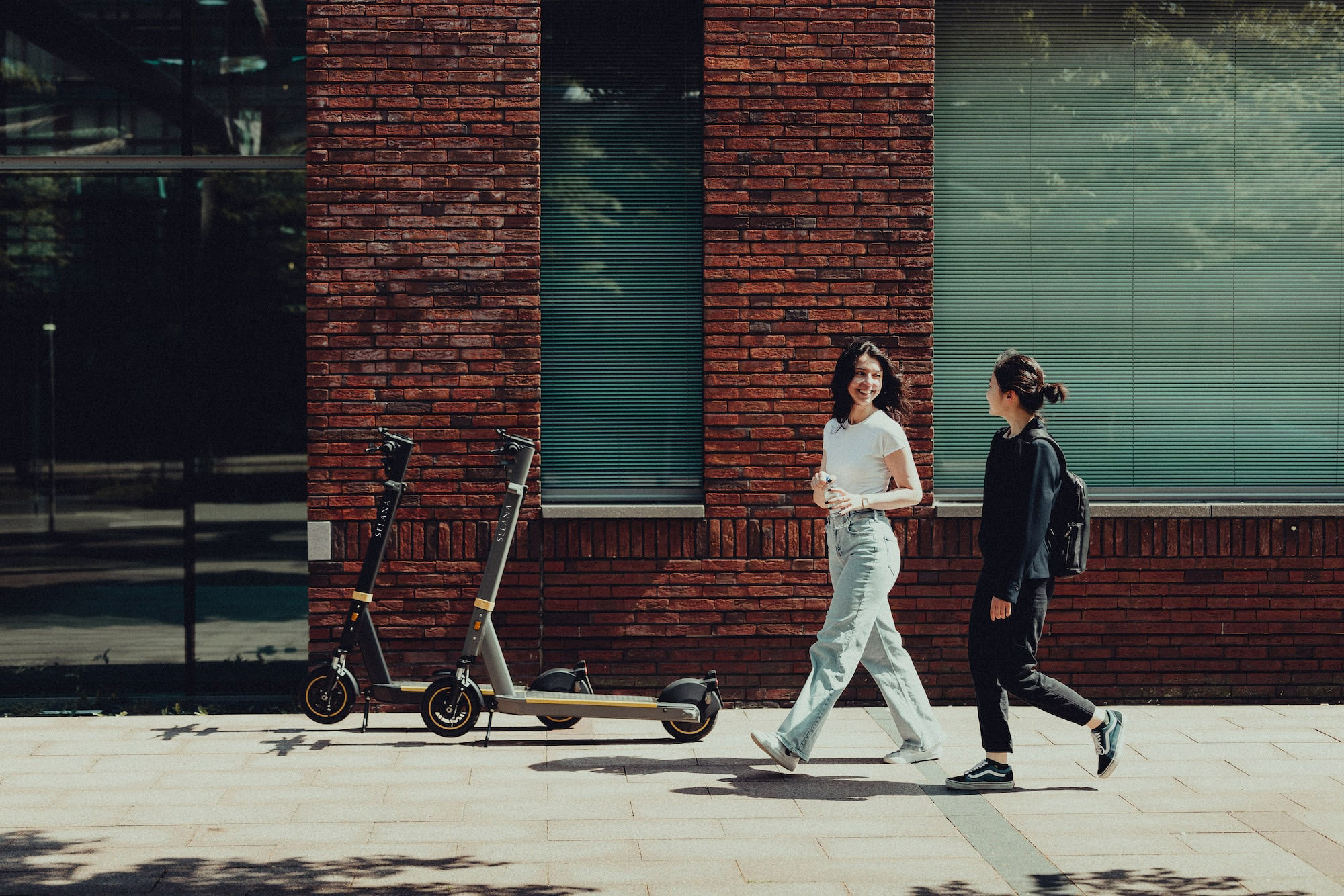 Two SELANA e-scooters parked by a brick facade, two women walking past