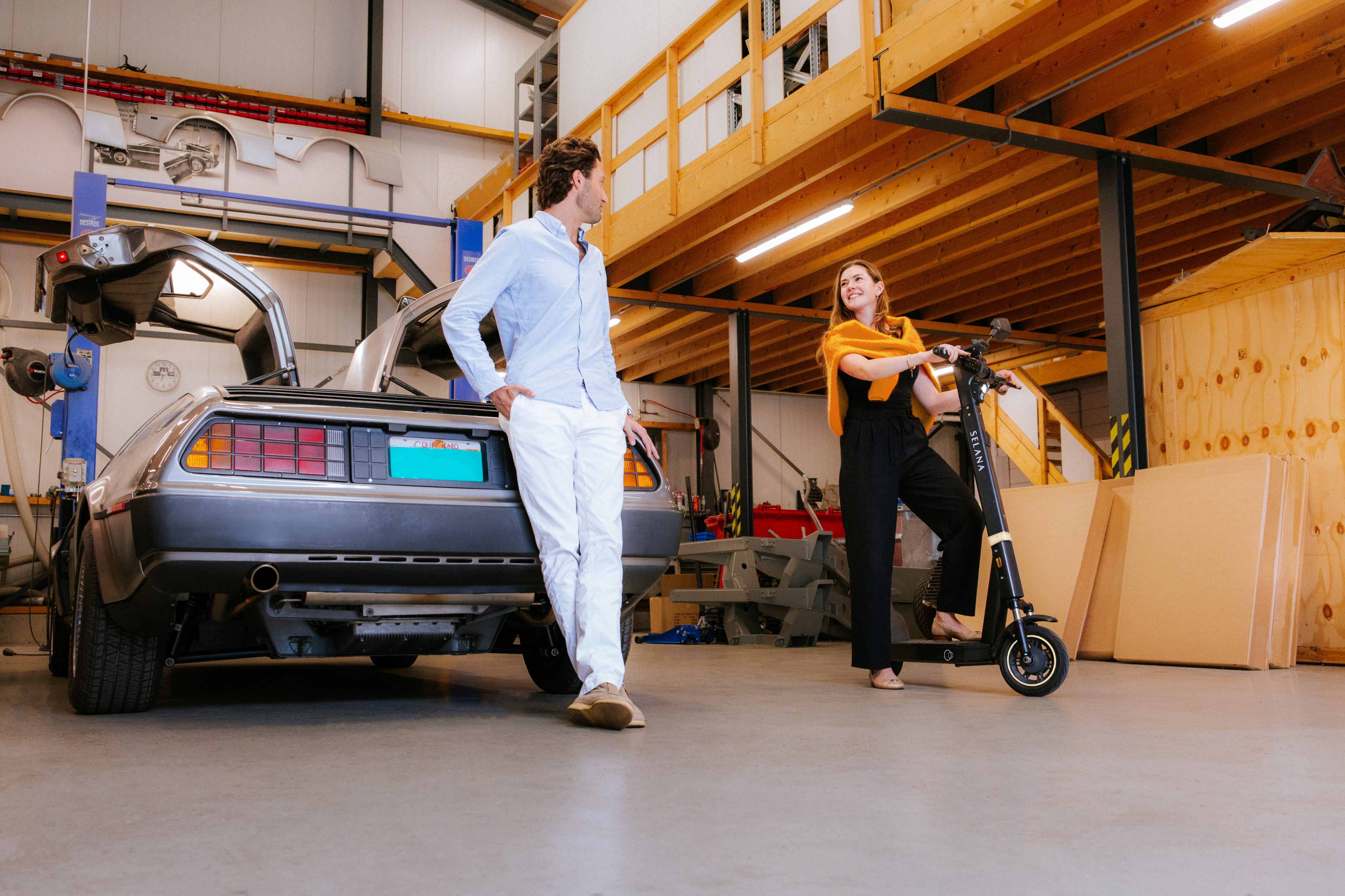 Two people with SELANA e-scooters in a garage next to a DeLorean